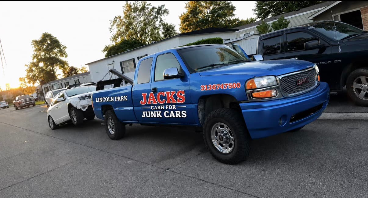 Blue GMC tow truck for Jacks Cash for Junk Cars towing a damaged white car.